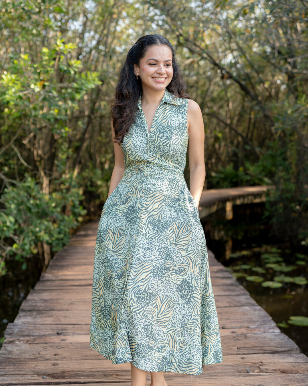 Woman in a green patterned dress standing on a wooden bridge with trees in the background