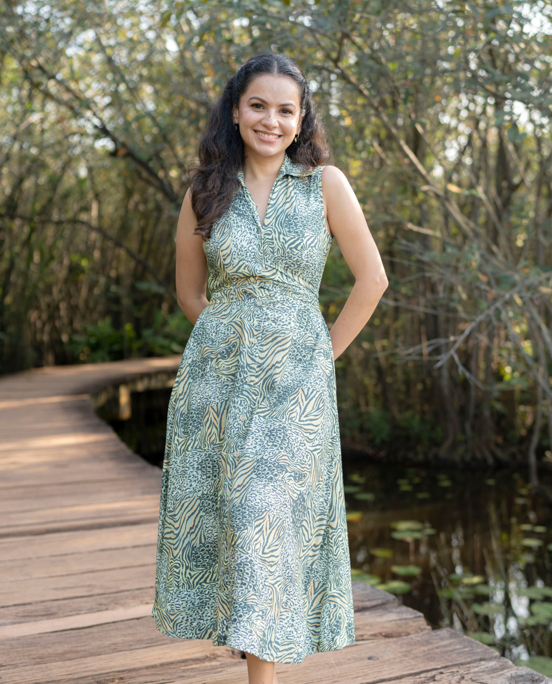 Woman in a green patterned dress standing on a wooden bridge with trees and water in the background